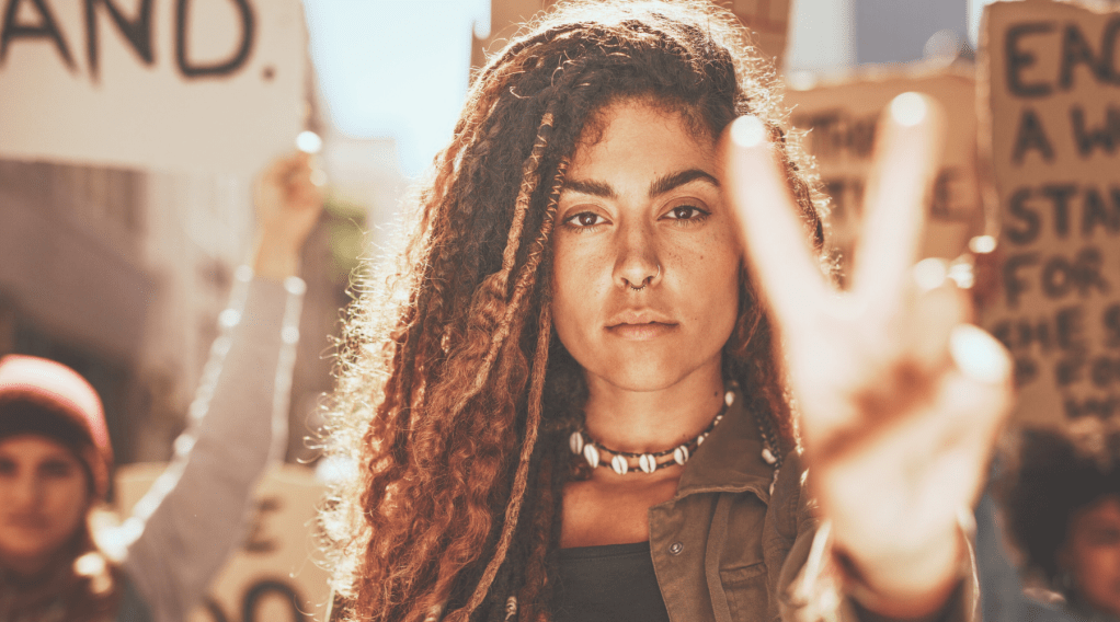 A woman stands with a crowd protesting while facing the camera and holding up two fingers representing the sign for peace